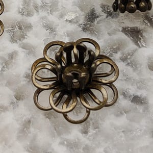 Può includere: Un cappuccio per perline a forma di fiore in metallo color bronzo con una coppa centrale e petali ad anello. Il cappuccio per perline è progettato per la creazione di gioielli e l'artigianato. Il cappuccio per perline è mostrato su uno sfondo marmorizzato bianco e grigio.