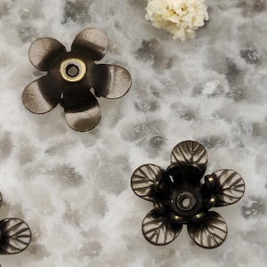 Può includere: Primo piano di diverse perline antiche a forma di fiore in bronzo. Ogni perlina ha cinque petali e un foro centrale, con un centro color oro. Le perline sono disposte su una superficie bianca e grigia simile al marmo.