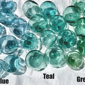 Puede incluir: Una colección de boyas de vidrio en varios tonos de azul, verde azulado y verde. Las boyas son redondas y tienen un acabado liso y brillante. Están dispuestas en un patrón aleatorio sobre una superficie blanca.
