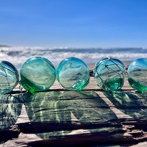 May include: Five spherical glass fishing floats in shades of translucent aqua and seafoam green. Some of the glass orbs are wrapped in thin, light-colored netting, while others are smooth and clear, reflecting the bright sunlight.