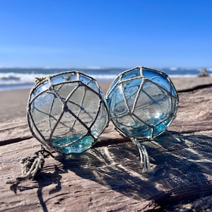 May include: Two spherical, translucent light blue glass fishing floats are encased in a decorative, hand-knotted beige rope netting. The glass spheres feature a smooth, reflective surface and are positioned side by side on a textured surface.