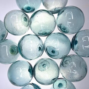 May include: A collection of twelve spherical glass floats in a pale blue-green hue. Each orb features a distinct circular pontil mark, indicating its handmade origin. The glass surfaces are smooth, translucent, and contain small air bubbles.