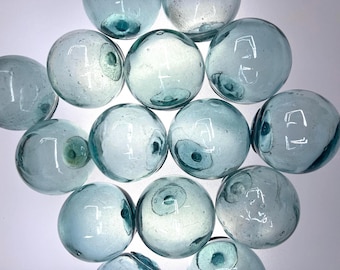 Puede incluir: Una colección de doce flotadores de vidrio esféricos en un tono azul verdoso pálido. Cada orbe presenta una marca de pontil circular distintiva, lo que indica su origen artesanal. Las superficies de vidrio son lisas, translúcidas y contienen pequeñas burbujas de aire.