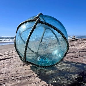 May include: A spherical, translucent light blue glass fishing float encased in a dark, weathered rope netting. The glass surface features visible air bubbles and a smooth texture, resting on a textured wooden surface near the ocean.