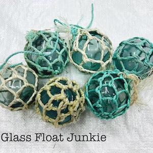 Pode incluir: Seis boias de pesca de vidro com um padrão de rede branco e verde. As boias são redondas e têm um tom azul claro. O texto "Glass Float Junkie" está impresso abaixo das boias.
