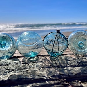 May include: Four spherical glass floats in shades of light blue and aqua. Three of the glass spheres are smooth, while one features a dark, thin netting wrapped around its exterior. The glass surfaces are translucent and contain small air bubbles.