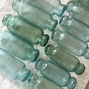 Könnte beinhalten: Eine Sammlung von 10 aquablauen Glas-Fischschwimmern, auch bekannt als "Netzschwimmer", mit abgerundeten Enden. Die Schwimmer sind in einem zufälligen Muster auf einer weißen Oberfläche angeordnet.