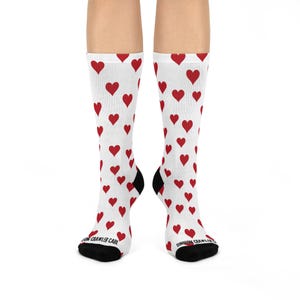 Puede incluir: Calcetines blancos con un patrón de corazones rojos. Los calcetines tienen talones y dedos negros. El texto "DUNGEON CRAWLER CARL" está impreso en la parte inferior de los calcetines.