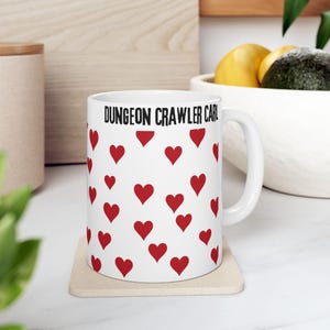 May include: White ceramic mug with red heart pattern and the text "DUNGEON CRAWLER CARL" in black. The mug is on a beige coaster. The mug has a white handle.