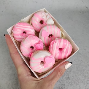 May include: A box of six pink and white donuts with various toppings, including pink icing swirls, heart-shaped sprinkles, and raspberry-shaped candies. The donuts are arranged in a white box with a light brown filling.