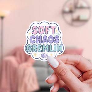 May include: A sticker with the words "SOFT CHAOS GREMLIN" in a pastel color scheme. The text is inside a cloud-shaped border with stars and a small design at the bottom. The sticker is held up by a person.