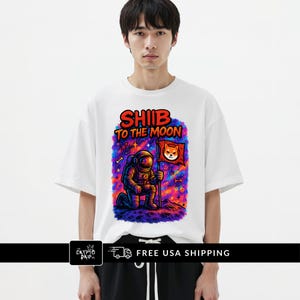 May include: White t-shirt featuring a colorful graphic of an astronaut on a planet with the text "SHIB TO THE MOON." The astronaut is holding a flag with a Shiba Inu dog. The shirt has a crew neck and short sleeves.