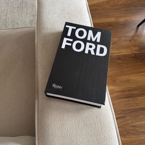 May include: A black book with the words "TOM FORD" in large white capital letters. The book is resting on a beige upholstered armrest. The book's spine reads "Rizzoli".