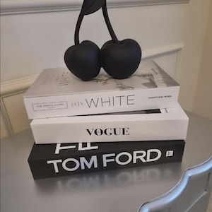 May include: A matte black cherry sculpture with a single leaf sits atop a stack of three books. The top book is titled WHITE by Christiana Nora, the middle book is titled VOGUE, and the bottom book is titled TOM FORD with a small TF logo.