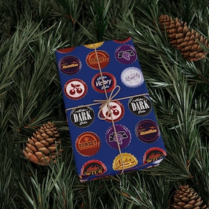 Puede incluir: Un regalo envuelto con un fondo azul que presenta diseños circulares con textos como "Nuka Dark Cola" y "Sunset Sarsaparilla". El regalo está atado con hilo y colocado sobre un fondo de agujas de pino y piñas.
