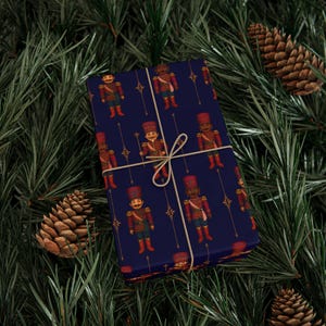 May include: A wrapped gift with a navy blue background and a repeating pattern of toy soldiers in red, gold, and green. The gift is tied with twine and set against a backdrop of pine needles and pine cones.