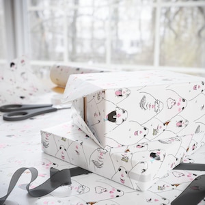Puede incluir: Dos cajas de regalo envueltas en papel blanco con un patrón repetido de caras de animales de dibujos animados en negro y rosa. También se ven un rollo de papel de regalo a juego, unas tijeras y una cinta negra.