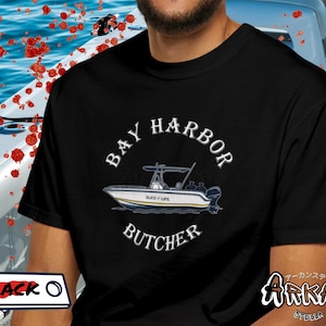 May include: Black t-shirt with a boat graphic and the text "BAY HARBOR BUTCHER". The boat is white and blue with the words "SLICE OF LIFE" on the side. The shirt has a crew neck and short sleeves.