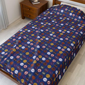 May include: A navy blue twin-size bedspread featuring a repeating pattern of colorful bottle caps. The caps are in various colors, including red, yellow, and white, with text and designs. The bedspread is on a wooden bed frame.