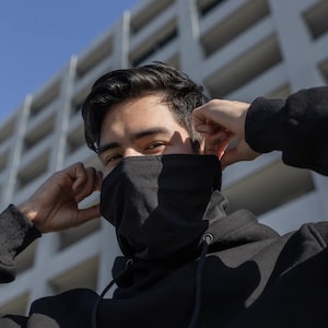 Puede incluir: Una persona con una mascarilla negra y una sudadera con capucha negra. La persona se está ajustando la mascarilla con ambas manos. El fondo presenta un edificio moderno y un cielo azul claro.