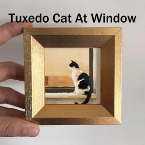 May include: A framed painting titled "Tuxedo Cat At Window" depicts a black and white cat sitting on a windowsill. The artwork is set in a gold-colored wooden frame. The cat is facing away from the viewer, gazing out the window.