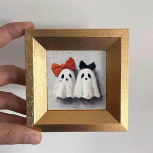 May include: A small square oil painting in a gold frame featuring two white ghost figures on a textured gray background. The left ghost wears a large orange bow, and the right ghost wears a small black bow. Both ghosts have black eyes and mouths.