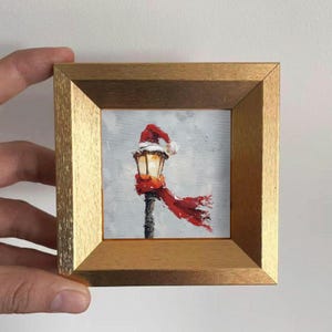 Può includere: Piccolo dipinto incorniciato raffigurante un lampione adornato da un cappello di Babbo Natale e una sciarpa rossa che sventola al vento. L'opera è incorniciata in una cornice di legno dorato, su sfondo grigio.