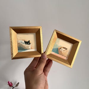May include: Two small square framed paintings. One features a black cat peeking out of a paper bag, the other a white cat. The frames are gold with a textured finish. The paintings have a soft, pastel color palette.