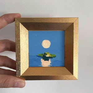 Peut inclure: Petite peinture encadrée représentant une grenouille sur un nénuphar sous la pleine lune. L'œuvre présente un ciel bleu, de l'eau et un cadre en bois doré. La grenouille est verte et jaune, et la lune est blanc crème.