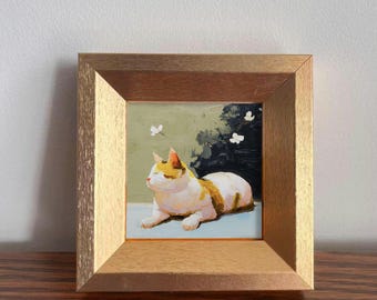 May include: A framed painting of a cat. The cat is white with orange and yellow markings, lying down with its eyes closed. The background features a light green wall with white flowers and dark green foliage. The frame is a light brown color.