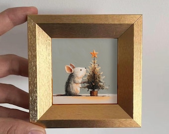 May include: A small square painting in a textured gold frame depicting a grey and white mouse standing next to a miniature Christmas tree topped with an orange star. The artwork features a light grey background and a white surface beneath the mouse.