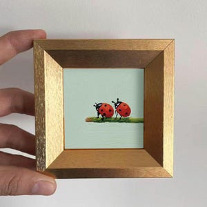 Puede incluir: Pequeña pintura enmarcada que representa dos mariquitas rojas con lunares negros posadas sobre un tallo verde, sobre un fondo azul claro. El marco cuadrado es dorado.