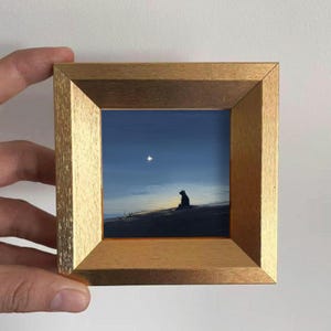 May include: A small framed painting depicts a silhouette of an animal on a hill under a starry night sky. The artwork is framed in a gold-colored wooden frame. The painting's color palette includes shades of blue, gold, and black.