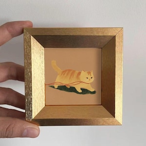 May include: A framed artwork featuring a cartoon cat with orange and cream stripes, walking on a green patch. The cat is depicted with a grumpy expression. The frame is gold-colored.
