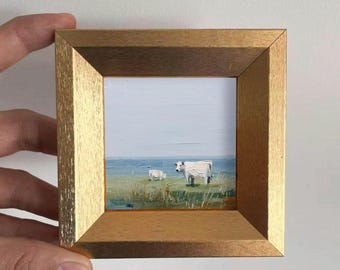 May include: A small, framed painting depicting two white cows in a field. The artwork features a gold-colored frame and a serene landscape with a blue sky and green grass. The cows are the focal point.
