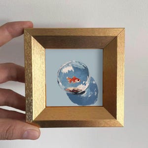 May include: A small square painting in a metallic gold frame featuring a single orange goldfish swimming inside a transparent glass bowl. The background is a solid light blue, and the bowl casts a dark blue shadow on the surface below.