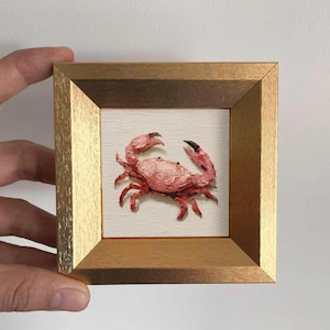 May include: A small, framed painting of a crab. The crab is painted in shades of pink and red with black claws. The frame is a light gold color, and the background is white.