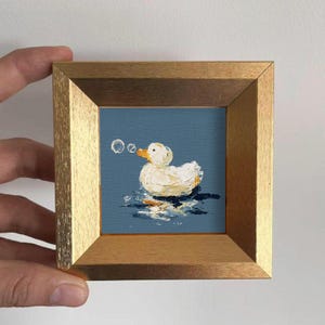 Può includere: Piccolo dipinto incorniciato di un'anatra bianca con becco arancione che galleggia su acqua blu, con bolle. La cornice è color oro chiaro.
