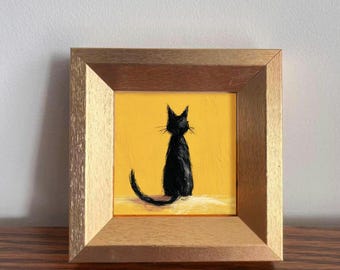May include: A framed painting of a black cat with a yellow background. The cat is depicted in a sitting position with its tail curved. The frame is a light brown color, and the painting is displayed on a wooden surface.