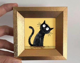 Puede incluir: Una obra de arte enmarcada con un gato negro caprichoso con grandes ojos blancos y bigotes, sobre un fondo amarillo. El gato está dibujado en un estilo de dibujos animados. El marco es de madera con acabado dorado.