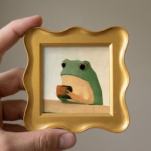 Könnte beinhalten: Ein kleines quadratisches Gemälde eines grünen Frosches, der eine braune Tasse hält, in einem dekorativen goldenen Rahmen mit gewelltem Rand. Der Frosch ist vor einem cremefarbenen Hintergrund dargestellt und stützt seine Arme auf eine helle Fläche.