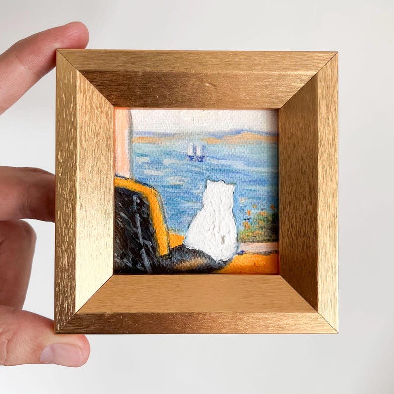 May include: Small framed painting featuring a white cat gazing out at a blue ocean scene with a sailboat. The artwork is set within a gold-colored wooden frame, creating a charming, miniature artwork.