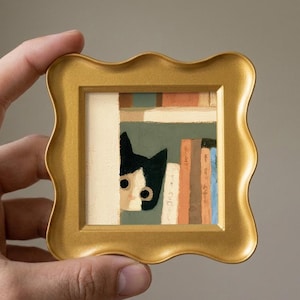 May include: A small square painting in a gold scalloped frame depicting a black and white cat peeking from behind a white wall next to a bookshelf with various colored books. The artwork features a flat, illustrative style with visible brushstrokes.