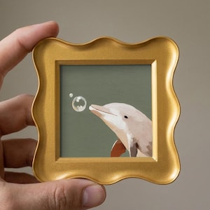 May include: A small square painting of a light pink dolphin blowing a bubble, set within a decorative gold-colored frame with a wavy, scalloped edge. The background of the painting is a solid muted olive green color.
