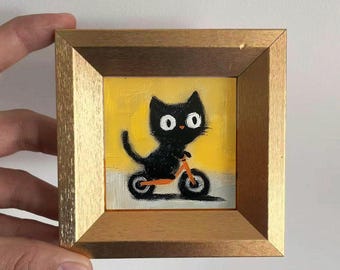 Puede incluir: Pintura enmarcada de un gato negro montando en bicicleta naranja sobre un fondo amarillo. El gato tiene grandes ojos blancos y está representado en un estilo de dibujos animados. El marco es de madera con un acento dorado.