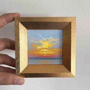 Puede incluir: Pequeña pintura al óleo de una puesta de sol sobre el mar, enmarcada en un marco de madera dorada. La obra presenta una vibrante paleta de amarillos, naranjas y rojos en el cielo, con un mar tranquilo azul-gris debajo. El marco es sostenido por una mano.