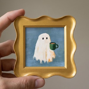 May include: A small square painting in a gold scalloped frame featuring a white ghost figure with two black eyes holding a dark green mug filled with a light brown beverage against a solid blue background. The artwork is painted with visible brushstrokes.