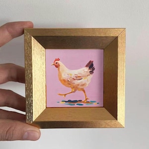 May include: A framed miniature painting of a chicken walking on a pink background. The chicken is white and orange with a red comb and a dark brown tail. The frame is gold-colored.