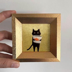 Puede incluir: Obra de arte enmarcada que presenta un gato negro sosteniendo un recipiente blanco con un pez naranja. El gato tiene ojos redondos y una larga cola. El fondo es dorado y el marco es de madera.