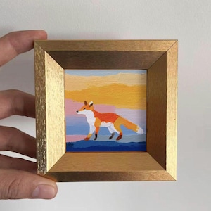 May include: A framed miniature painting of a fox. The fox is depicted in orange, white, and black, walking across a blue surface. The background features a colorful sunset with hues of pink, yellow, and blue. The frame is a light gold color.